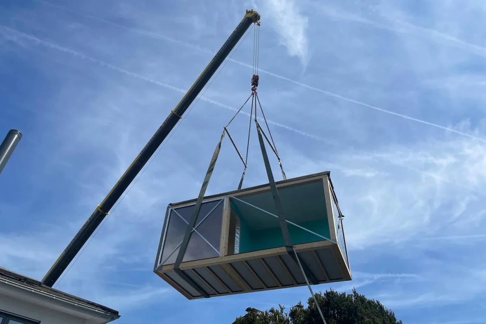 Ein fertig gebauter Cube hängt am Haken eines Autokrans und wird über das Grundstück gesetzt.