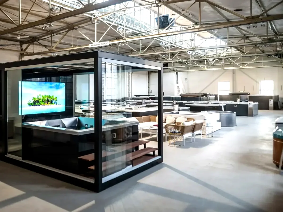 REINHART Spa Cube in einer Ausstellungshalle mit Whirlpool, Holzstufen und großem Bildschirm hinter Glas.